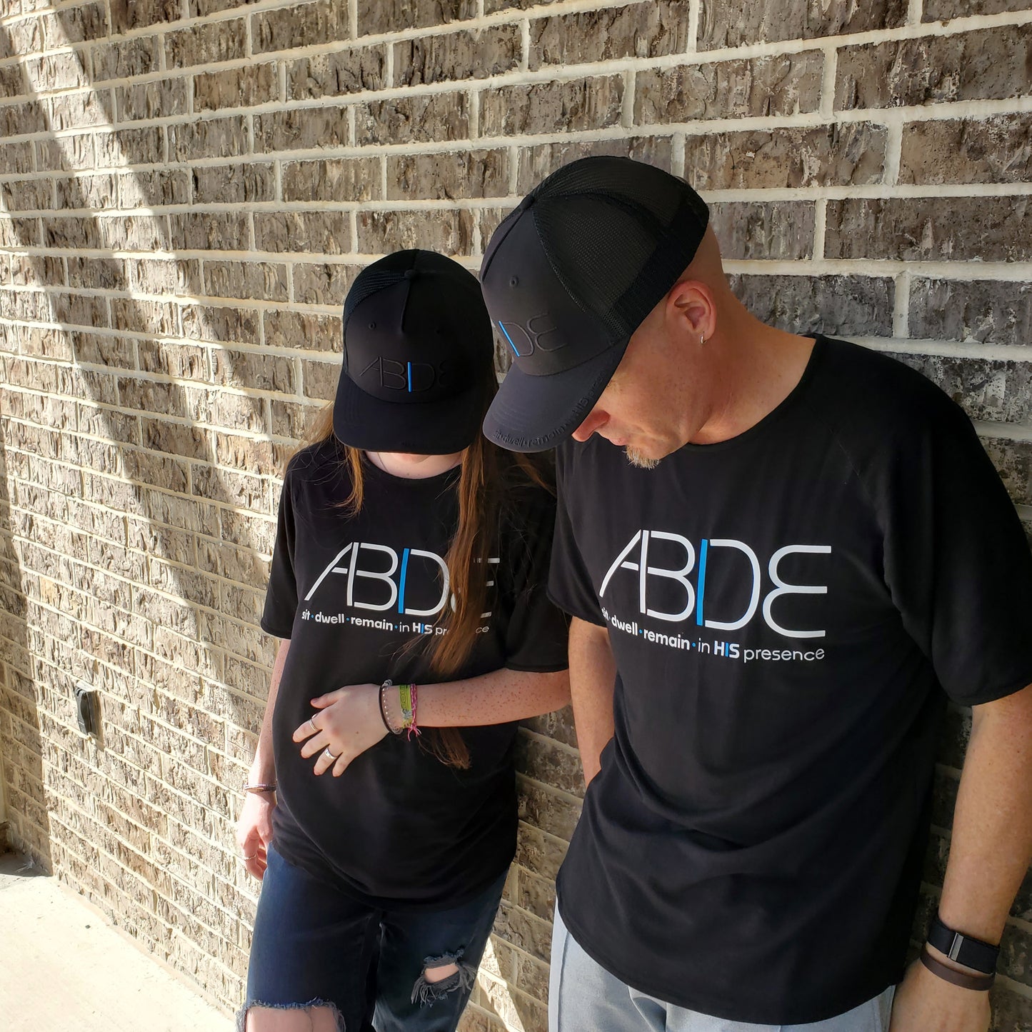 A father and daughter wearing matching midnight black "Abide" bamboo t-shirts and "Abide" blackout trucker hats from Now Found Apparel, offering Christian apparel