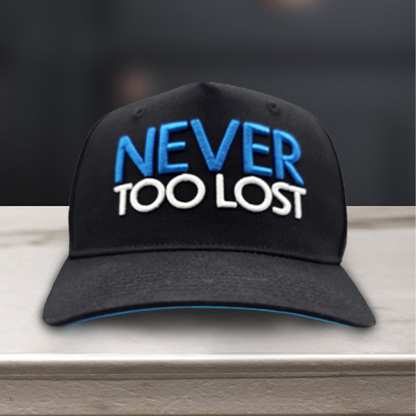 Black cap with 'NEVER TOO LOST' text on front and 'TO BE FOUND' on the back sitting on a marble surface