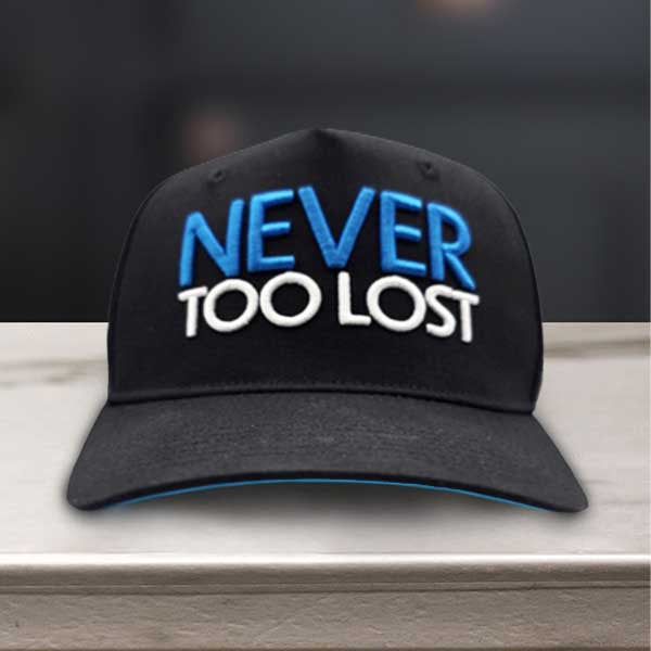 Black cap with 'NEVER TOO LOST' text on front and 'TO BE FOUND' on the back sitting on a marble surface