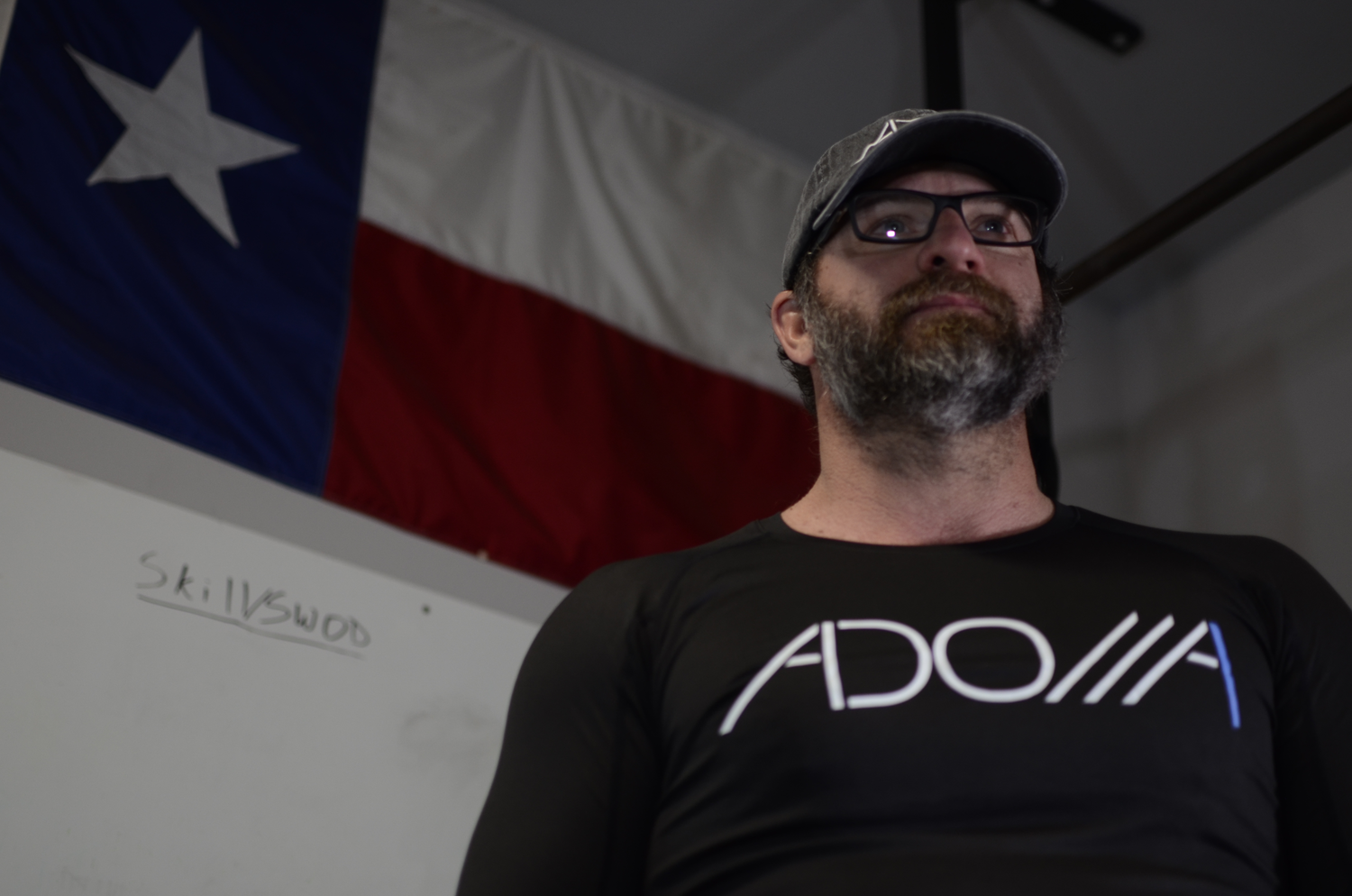 Now Found Model standing in a gym with a Texas flag in the background wearing Now Found's ADONAI Long-Sleeve Fitness Tee