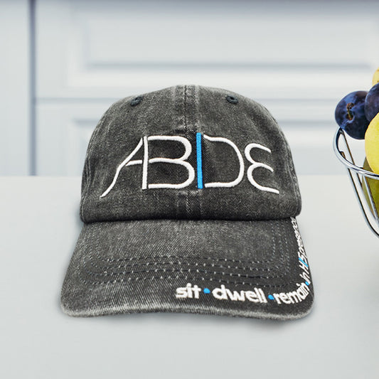 Charcoal gray cap with 'ABDE' sit - dwell - remain in His presence on a white counter with fruit in the background