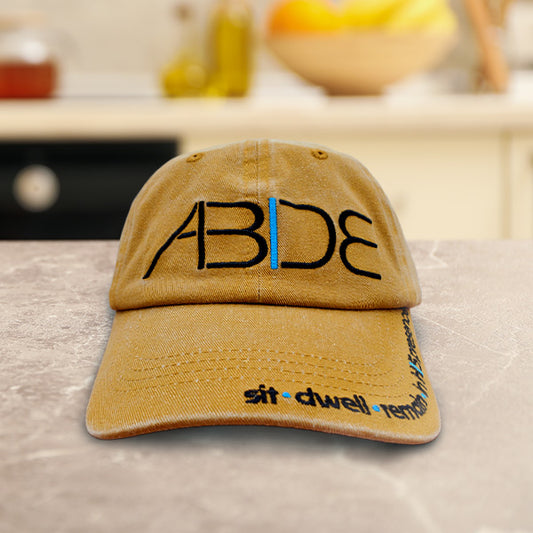 Mustard Yellow cap with 'ABDE sit - dwell - remain in His presence' on a countertop