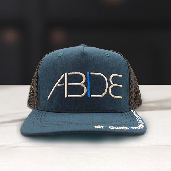 Blue Christian Trucker Hat with 'ABIDE' sit - dwell - remain in His Presence logo on a wooden surface