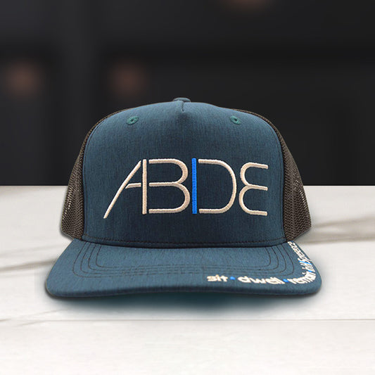 Blue Christian Trucker Hat with 'ABIDE' sit - dwell - remain in His Presence logo on a wooden surface