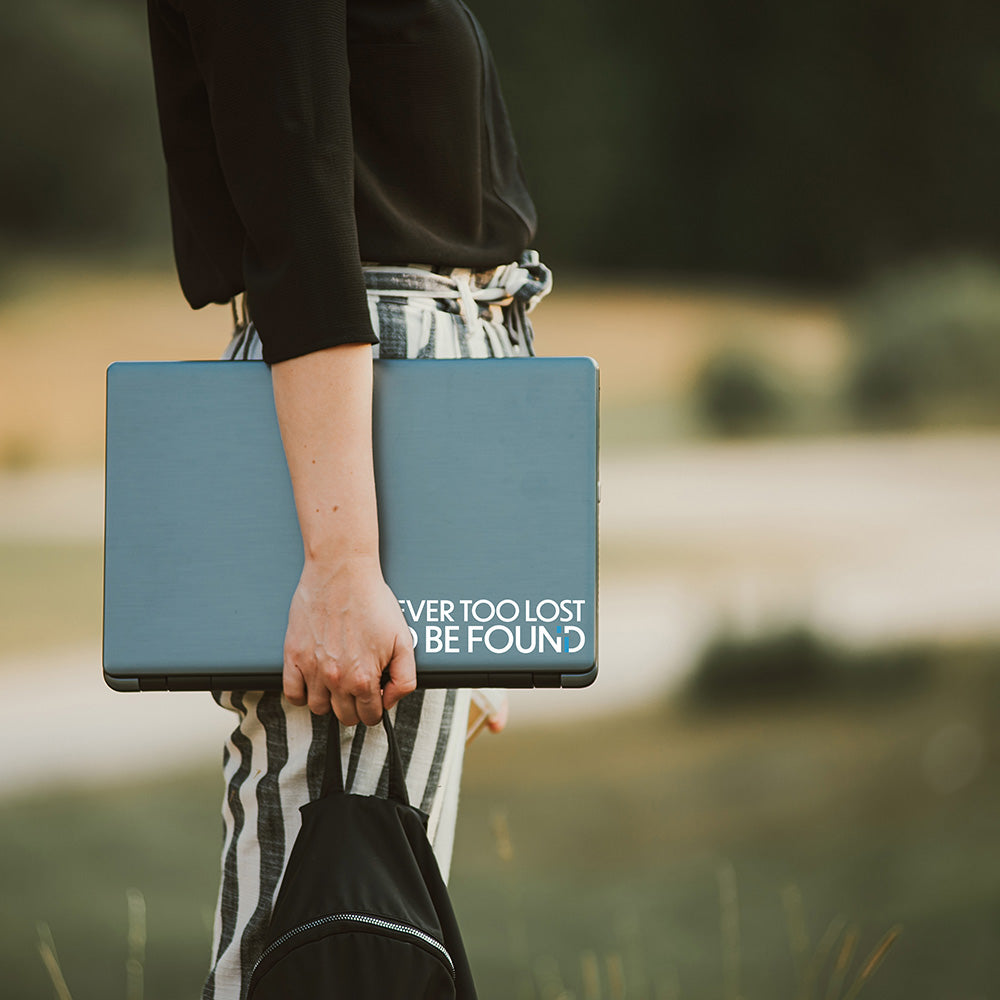 student standing in park with laptop in hand with Never Too Lost To Be Found sticker
