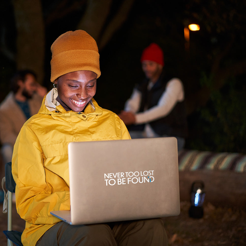young woman camping working on laptop with Never Too Lost To Be Found vinyl sticker on it
